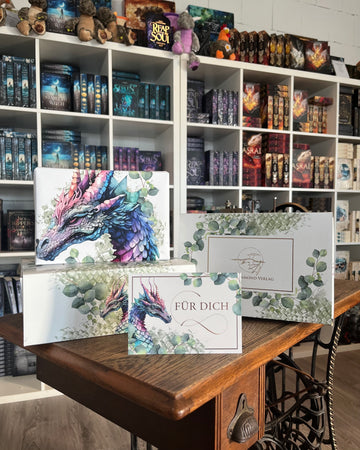 Das Bild zeigt auf einer alten Nähmaschine und vor den Bücherregalen im Drachennest eine Drachenpostbox von oben, eine Drachenpostbox von der Seite, ein in ein Drachen-Poster verpacktes Buch und eine Postkarte mit einem Drachen und dem Schriftzug "Für Dich". Die Hintergründe von Box, Poster und Karte sind weiß, der je darauf gezeigte Drache ist in Schattierungen der Farben Violett, Lila und Grün gestaltet. Schuppen und Eukalyptus-Blätter ziehen sich zusätzlich über Box, Poster und Karte.